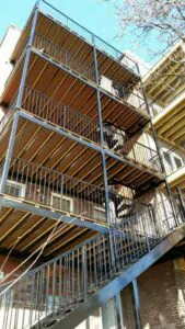 Four-story metal fire escape with stairs, iron balconies, and railings attached to a brick building, seen from ground level.